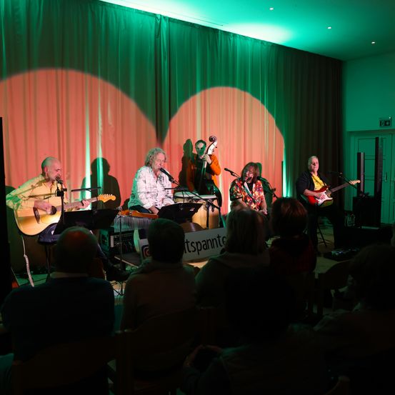 Eine Gruppe von Musikern tritt auf der Bühne vor einem Publikum auf. Einer spielt Gitarre, ein anderer Cello, und zwei singen. Die Bühne ist mit grünen und orangefarbenen Vorhängen dekoriert.