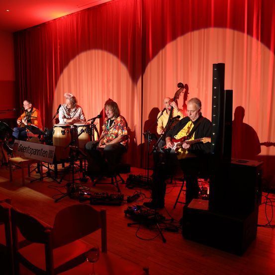 Eine Band tritt auf einer Bühne mit einem roten Vorhang im Hintergrund auf. Die Musiker umfassen einen Gitarristen, einen Bassisten, einen Perkussionisten und zwei Sänger.