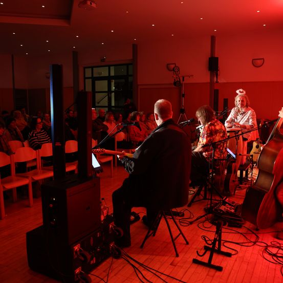 Eine Veranstaltung in einem Gebäude mit einer Band auf der Bühne. Der Leadsänger sitzt auf einem Stuhl mit einer Gitarre. Das Publikum sitzt auf Stühlen. Die Bühne ist mit roten Lichtern beleuchtet.
