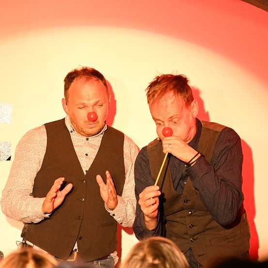 Zwei Männer mit braunen Westen und roten Clownsnasen treten auf der Bühne vor einem Publikum auf. Ein Mann bläst einen gelben Stock.