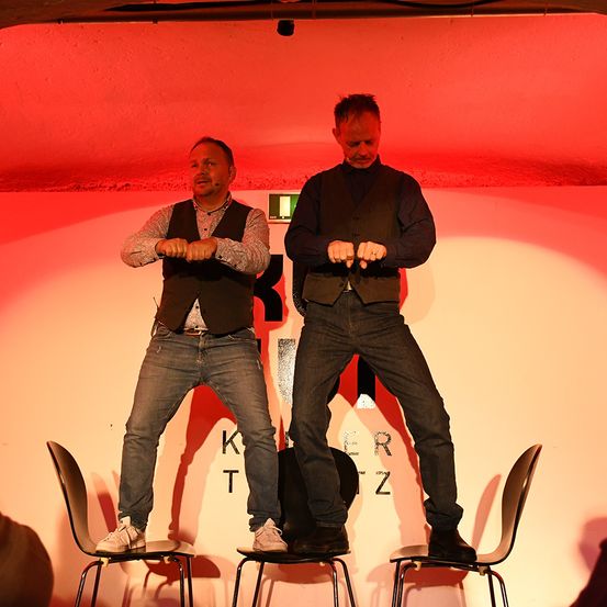 Zwei Männer auf der Bühne in Jeans und Weste stehen auf Stühlen und führen vermutlich eine Comedy-Show auf. Der Hintergrund zeigt eine rote Wand mit weißen Buchstaben, die 'K T Z' lesen.