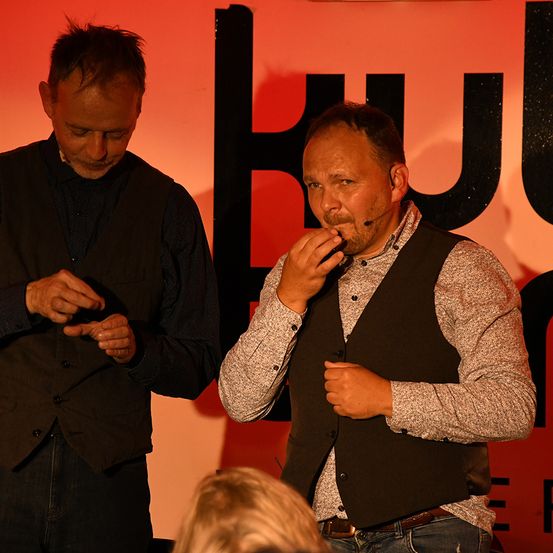 Zwei Männer auf der Bühne, einer gestikuliert, der andere isst, mit dem Kopf einer Frau im Vordergrund. Dahinter ein roter Hintergrund mit schwarzem Text.