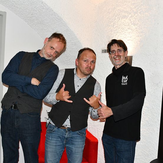 Drei Männer stehen in einem Raum, zwei machen Gesten. Der Mann auf der linken Seite hat die Arme verschränkt, der Mann in der Mitte macht Handgesten und der Mann auf der rechten Seite trägt eine Brille und ein schwarzes Shirt mit einem weißen Logo. Hinter ihnen steht ein roter Stuhl und eine weiße Wand.