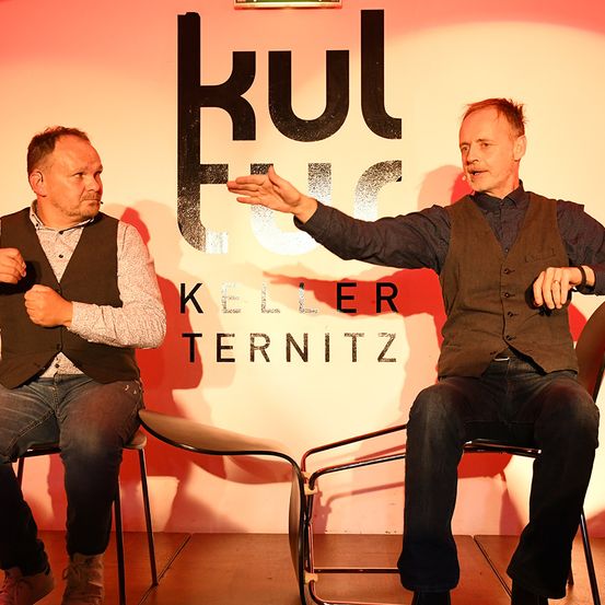 Zwei Männer sitzen vor einer rosa Wand mit dem Wort 'Kul' in schwarzen Buchstaben. Ein Mann zeigt mit der Hand, während der andere ihn ansieht.