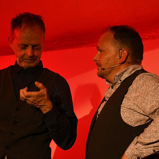 Zwei Männer stehen unter rotem Licht, einer mit Mikrofon, beide in schwarzen Westen. Der Mann auf der linken Seite gestikuliert mit den Händen.