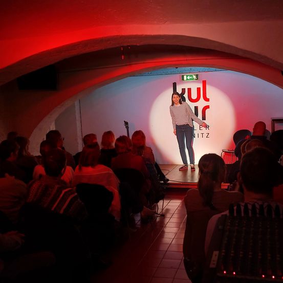 Eine Frau steht auf der Bühne unter einer Lampe und tritt vor einem Publikum auf, das in einem dunkel beleuchteten Raum mit roter Beleuchtung sitzt.