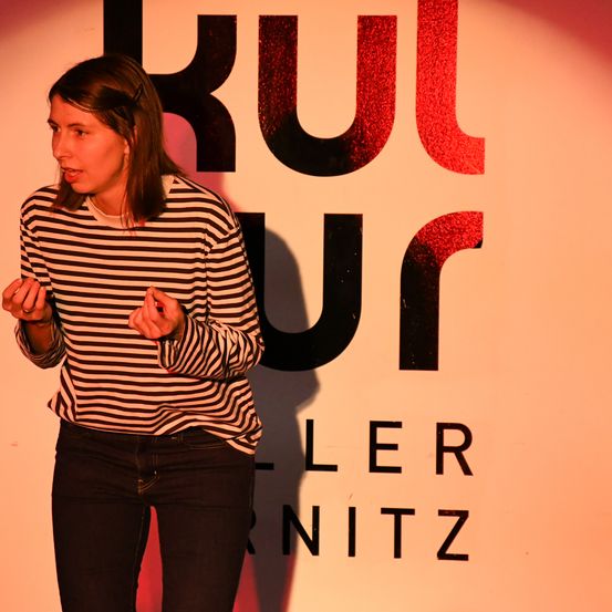 Eine Frau in einem gestreiften Shirt steht vor einer Wand mit roten Buchstaben, die 'kul ur ler nitz' lesen. Sie gestikuliert mit den Händen, während sie spricht.