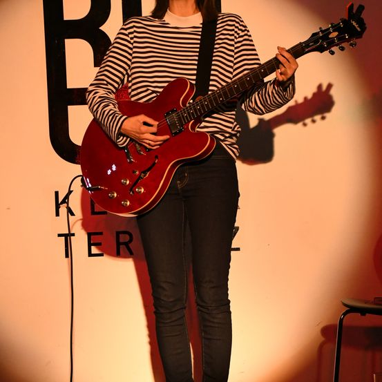 Eine Frau spielt eine rote Gitarre auf der Bühne unter Bühnenbeleuchtung. Sie trägt ein gestreiftes Hemd und schwarze Jeans. Hinter ihr ist eine Wand mit dem Wort 'Kelter'.