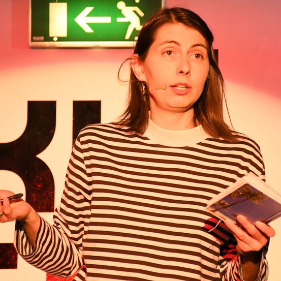 Eine Frau mit Mikrofon spricht und hält ein Buch vor sich vor einer rosa Wand mit einem Schild für einen Ausgang.