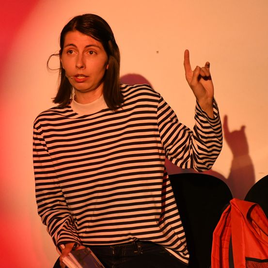 Eine Frau in einem gestreiften Shirt sitzt auf einem Stuhl, macht ein Friedenszeichen und hält ein Buch. Ein roter Rucksack ist neben ihr.