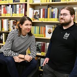 Eine Frau in einem gestreiften Shirt sitzt auf einem Hocker, während ein Mann in einem schwarzen Shirt neben ihr steht in einem Buchladen mit Bücherregalen voller Bücher im Hintergrund.