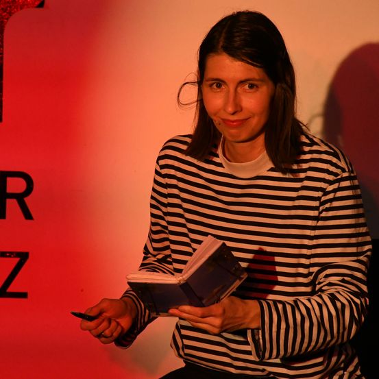 Eine Frau in einem schwarz-weiß gestreiften Shirt hält ein Buch, lächelt und liest, vor einem roten und weißen Hintergrund.