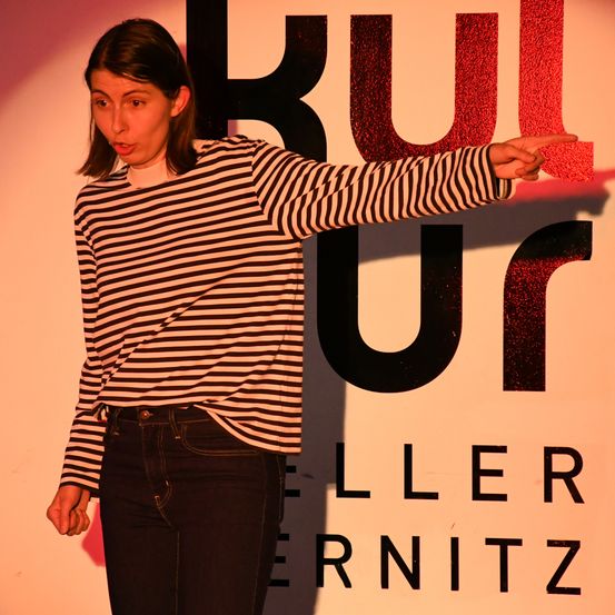 Eine Frau in einem gestreiften Shirt und Jeans steht vor einem Schild mit dem Wort 'KULTUR' in fetten Buchstaben. Sie zeigt mit der rechten Hand auf das Schild.