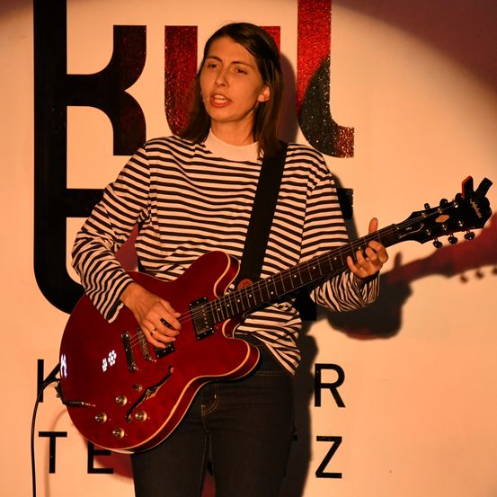 Eine Frau in einem gestreiften Shirt spielt eine rote E-Gitarre auf der Bühne. Der Hintergrund hat große Buchstaben, die 'BULLET' buchstabieren.