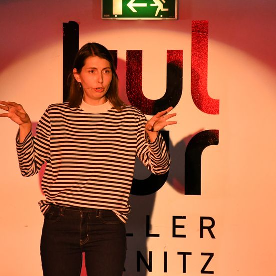 Eine Frau mit einem Mikrofon steht vor einer Wand mit roten und schwarzen Buchstaben, die 'uler' und 'er nitz' lesen. Sie trägt ein gestreiftes Hemd und Jeans.