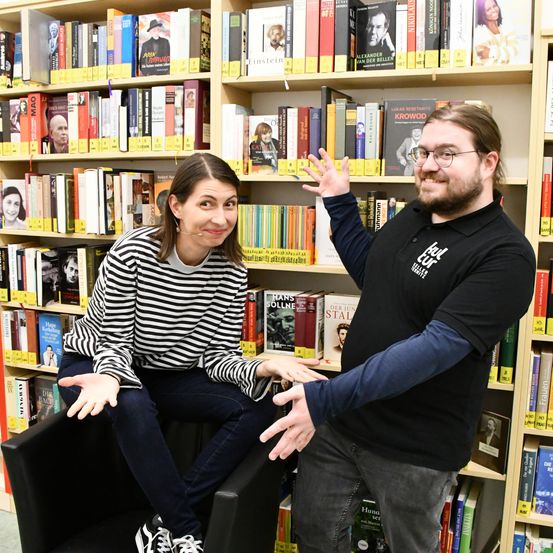 Ein Mann und eine Frau stehen vor einem Bücherregal voller Bücher. Der Mann trägt eine Brille und ein schwarzes T-Shirt mit weißem Text. Die Frau trägt ein gestreiftes Shirt und sitzt auf einem Stuhl. Sie lächeln beide und schauen in die Kamera.