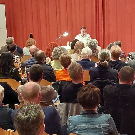 Ein Mann mit Brille sitzt an einem Schreibtisch und spricht eine Menschenmenge in einem Raum mit roten Vorhängen an. Das Publikum besteht aus Männern und Frauen verschiedenen Alters.