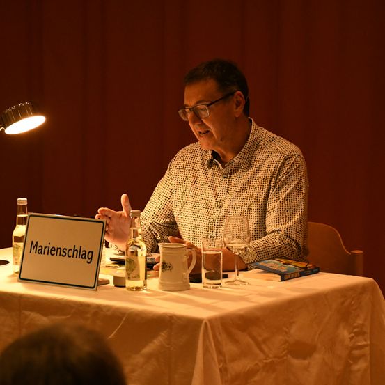 Ein Mann sitzt beleuchtet von einer Lampe an einem Tisch und spricht zu einem Publikum. Der Tisch hat ein Namensschild, Getränke und ein Buch.