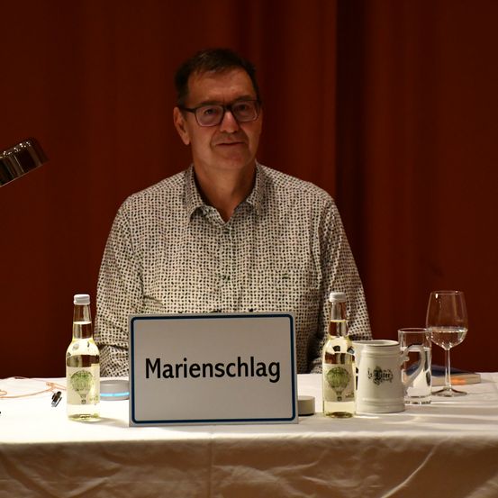 Ein Mann sitzt an einem Tisch mit einem Namensschild, das Marienschlag steht. Auf dem Tisch befinden sich Flaschen, Weingläser und Tassen. Eine Lampe ist hinter ihm positioniert.