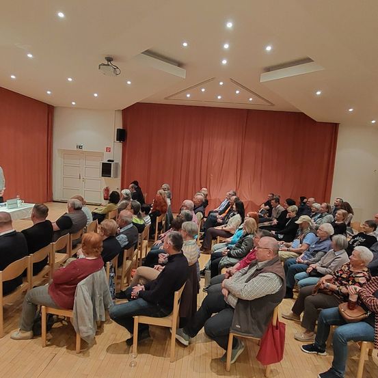Eine Gruppe von Menschen sitzt in einem Raum mit einer Bühne und hört einer Präsentation zu. Die Bühne hat einen Tisch und einen Projektor. Der Raum hat einen roten Vorhang und Scheinwerfer.