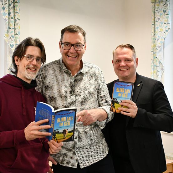 Drei Männer posieren für ein Foto in einem Raum. Der Mann auf der linken Seite hält ein Buch, der in der Mitte lächelt und der auf der rechten Seite hat eine Kamera.