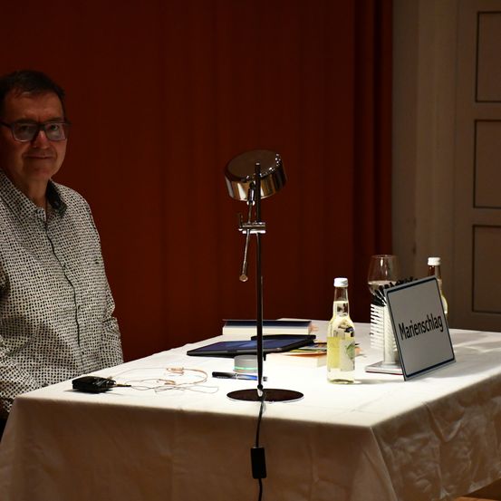 Ein Mann mit Brille steht vor einem Schreibtisch mit Lampe, Schild und verschiedenen Gegenständen. Er befindet sich in einem schwach beleuchteten Raum mit einem roten Vorhang und einer Tür hinter ihm.