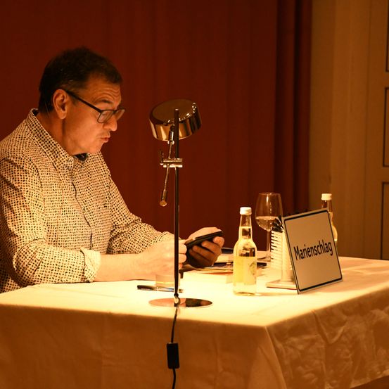 Ein Mann mit Brille sitzt an einem Tisch, benutzt ein Smartphone, mit einer Schreibtischlampe und Weingläsern. Ein Schild auf dem Tisch sagt 'Marienschalg.'