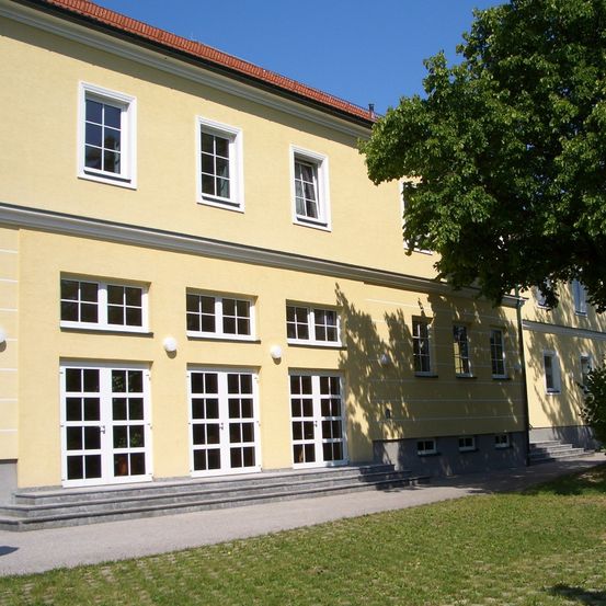 Der Außenbereich eines gelben Gebäudes mit weißen Verkleidungen und mehreren Fenstern ist zu sehen, neben dem ein Baum steht.
