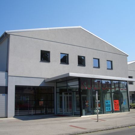 Ein modernes, graues Gebäude mit Glasfenstern und Türen und einem Schild, das Kulturhaus besagt. Das Gebäude hat ein dreieckiges Dach und steht an einem klaren Tag.