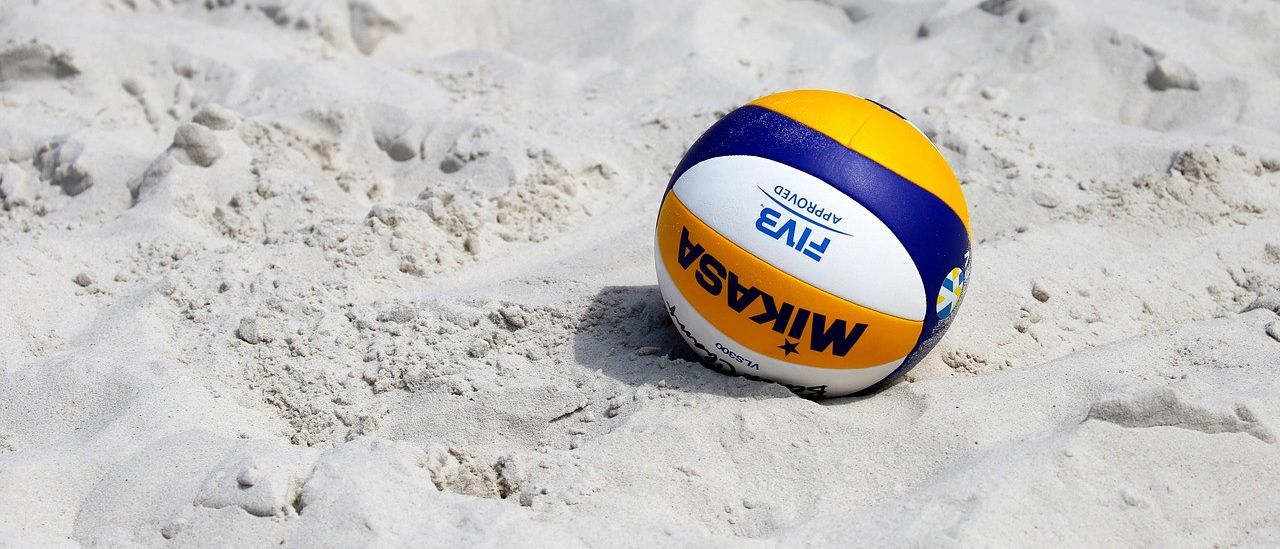 Ein Strandvolleyball liegt auf dem Sand und zeigt gelbe, blaue und weiße Farben mit den Worten 'Wilson' und 'FIVB' darauf geschrieben.