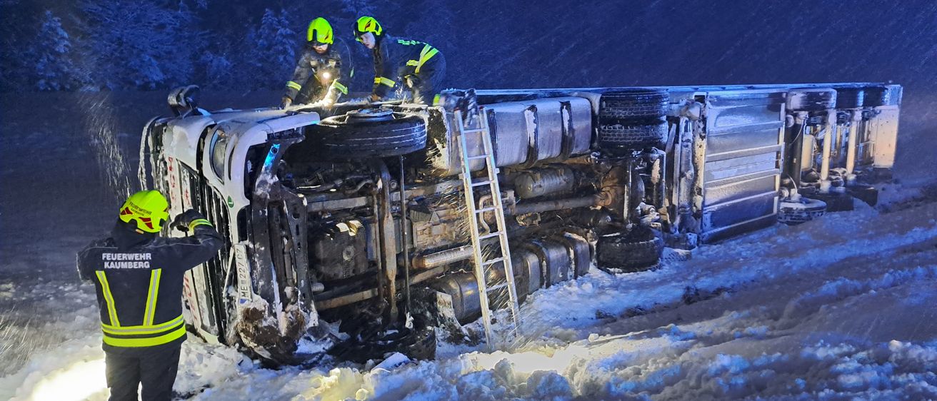 Zwei Feuerwehrleute in Helmen und Uniformen inspizieren einen beschädigten Lkw im Schnee. Eine Leiter ist gegen den Lkw gestellt.