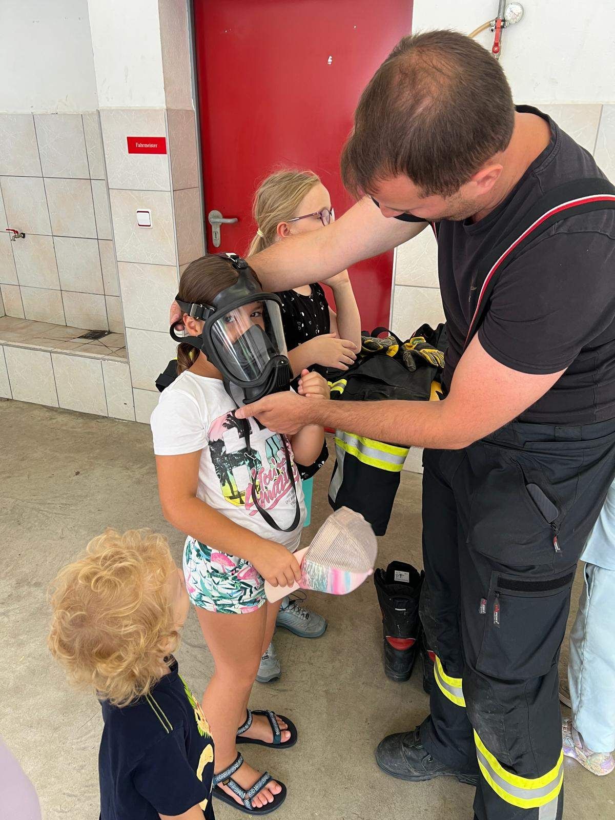 Ein Feuerwehrmann hilft einem kleinen Mädchen, eine Gasmaske anzulegen, während ein Kleinkind zuschaut. Dahinter beobachtet eine Frau mit Brille.