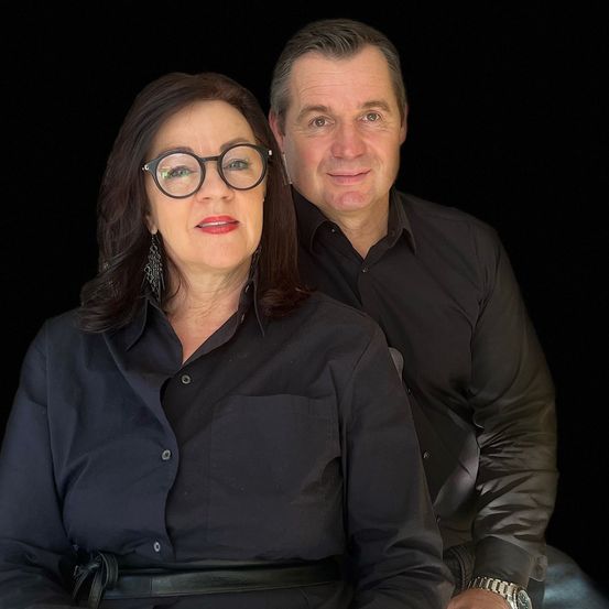 Ein Mann und eine Frau sitzen gemeinsam vor schwarzem Hintergrund, beide tragen schwarze Hemden. Die Frau trägt eine Brille und Ohrringe, der Mann hat eine Uhr an seinem linken Handgelenk. Sie scheinen beide für die Kamera zu lächeln.