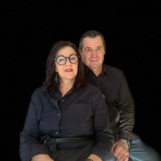 Ein Mann und eine Frau sitzen gemeinsam vor schwarzem Hintergrund, beide tragen schwarze Hemden. Die Frau trägt eine Brille und Ohrringe, der Mann hat eine Uhr an seinem linken Handgelenk. Sie scheinen beide für die Kamera zu lächeln.