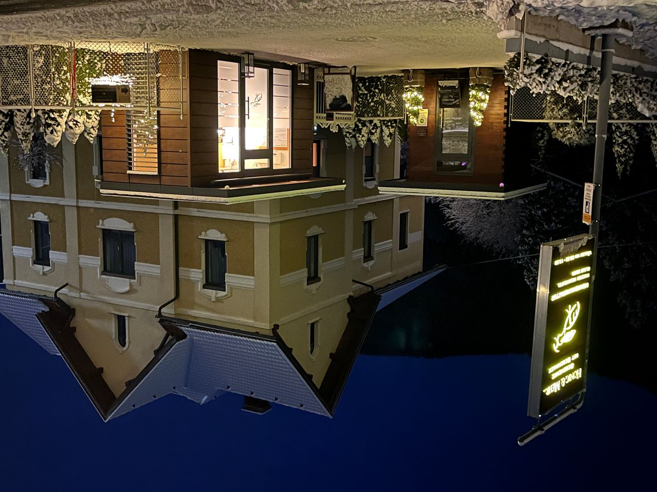 Eine Luftaufnahme eines Hauses, das sich im Wasser spiegelt, mit einem beleuchteten Fenster, Pflanzen und Schildern an der Seite.