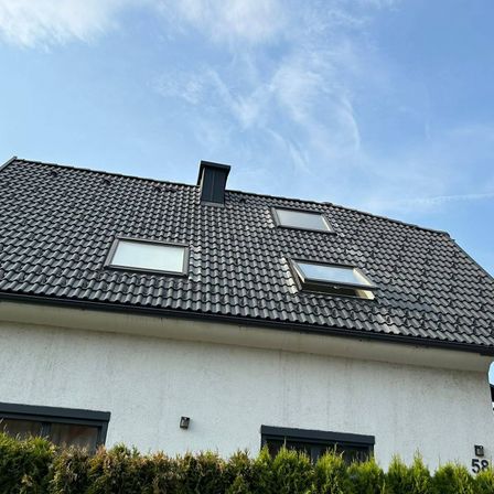 Ein weißes Haus mit grauem Dach hat drei Fenster, darunter zwei Dachfenster, unter einem blauen Himmel mit verstreuten Wolken.
