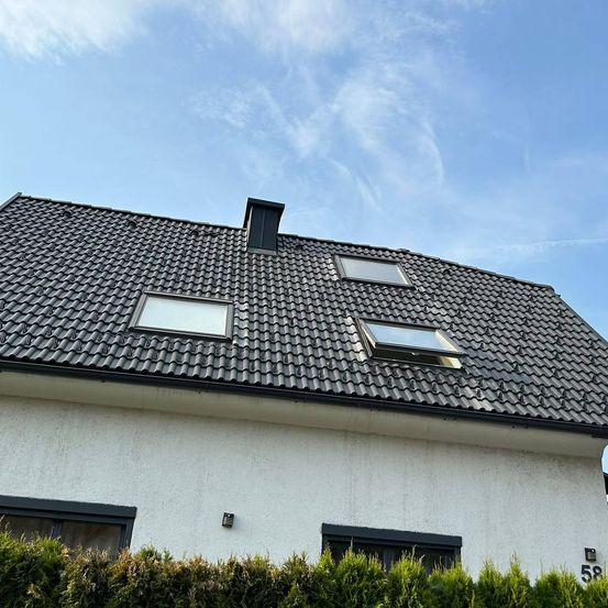 Ein weißes Haus mit grauem Dach hat drei Fenster, darunter zwei Dachfenster, unter einem blauen Himmel mit verstreuten Wolken.