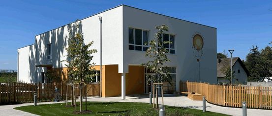 Ein modernes weißes Gebäude mit einem gelben Akzent und einem Logo an der Seite. Es hat mehrere Fenster und einen doppelten Türeingang. Davor befindet sich ein kleiner Garten mit einem Rasen, einigen Bäumen und einem Holzzaun.