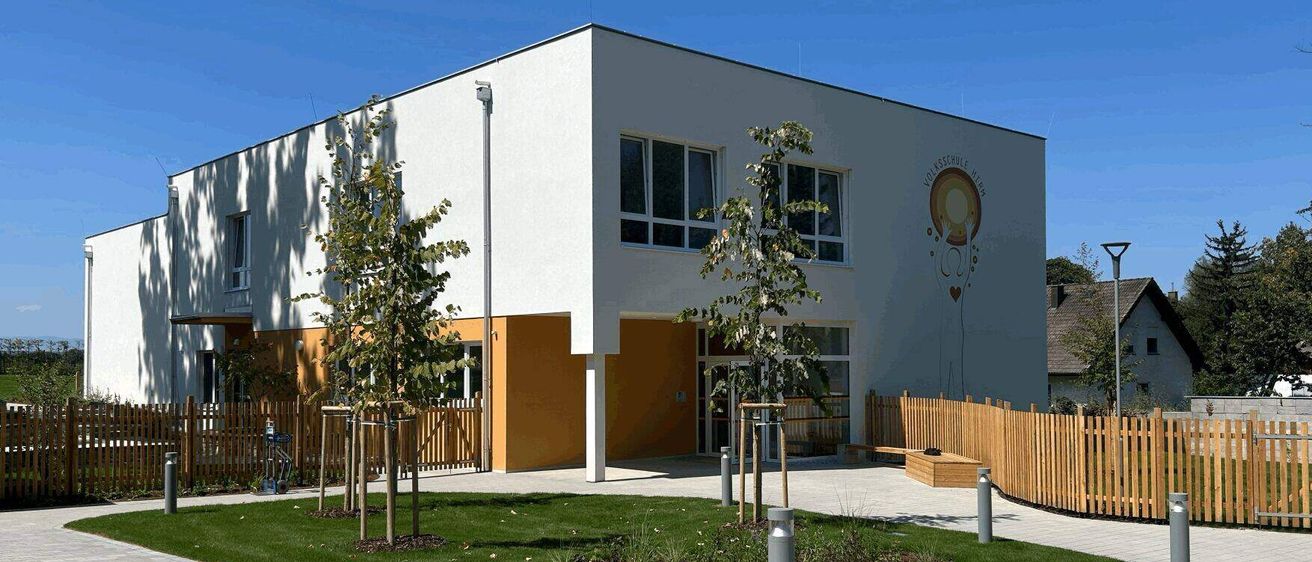 Ein modernes weißes Gebäude mit einem gelben Akzent und einem Logo an der Seite. Es hat mehrere Fenster und einen doppelten Türeingang. Davor befindet sich ein kleiner Garten mit einem Rasen, einigen Bäumen und einem Holzzaun.