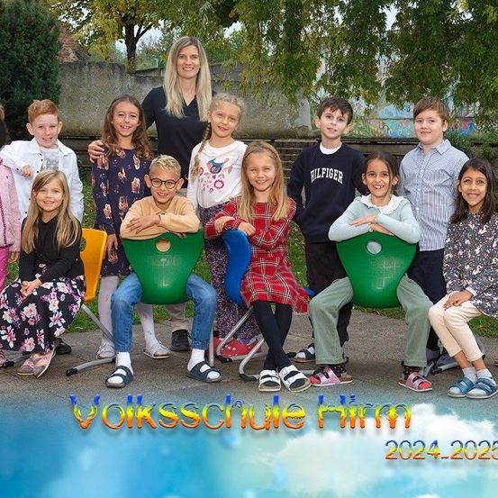 Eine Gruppe von Kindern und einer Frau steht draußen auf einem Betonweg mit Bäumen im Hintergrund. Sie posieren für ein Foto. Sie sitzen auf grünen Stühlen, und einige stehen dahinter. Der Text am unteren Rand lautet 'Volksschule Himm 2024-2025'.