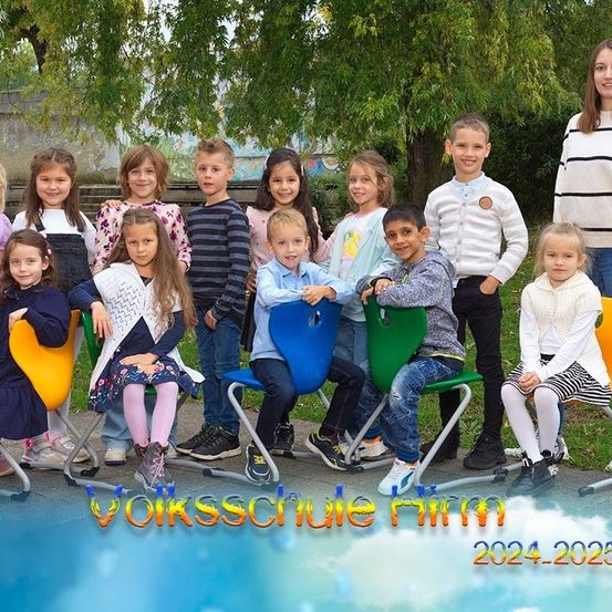 Eine Gruppe von Kindern und zwei Erwachsenen lächelt und posiert für ein Foto in einem Park, mit einem Baum und einer Graffitiwand im Hintergrund. Die Kinder sitzen auf bunten Stühlen, und das Foto hat einen Wasserzeichen mit dem Text 'Volksschule Himm 2024-2025'.