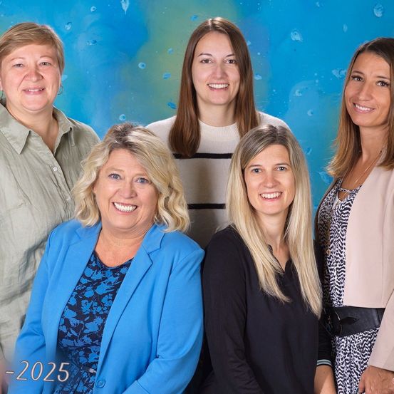 Fünf Frauen in Geschäftskleidung stehen vor einem blau-grünen Hintergrund. Sie alle lächeln für ein Gruppenfoto. Die Frau in der Mitte trägt einen weißen Pullover. Die Frau auf der linken Seite trägt einen blauen Blazer. Unter dem Bild steht das Jahr 2025 geschrieben.