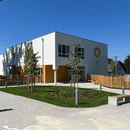 Ein modernes Gebäude mit weißen Wänden und gelben Akzenten steht an einem sonnigen Tag. Es hat große Fenster, ein Logo an der Wand und einen kleinen Garten davor.