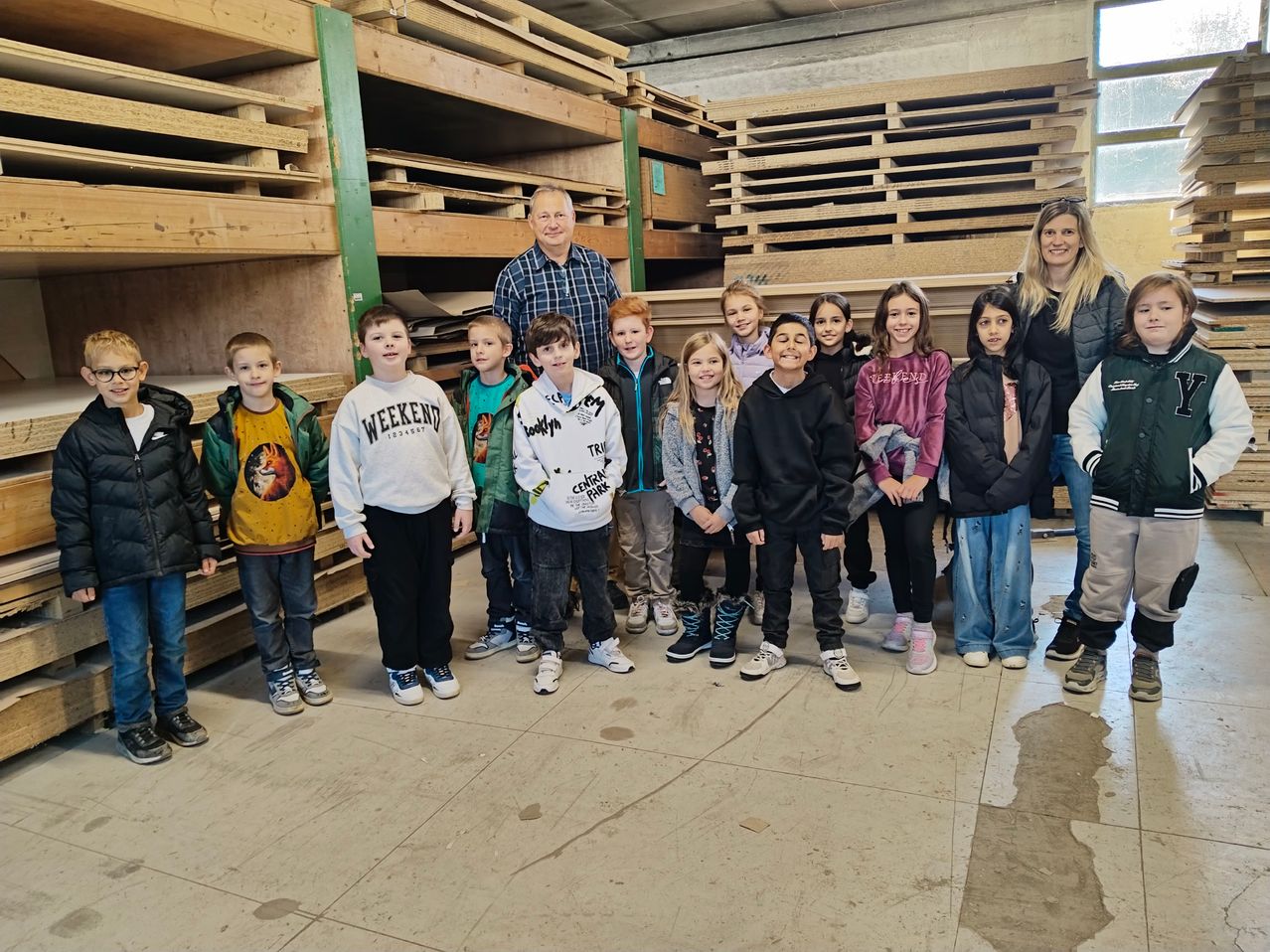 A group of children and two adults are standing in a room with wooden pallets stacked behind them. The children are wearing jackets, while the adults are wearing casual clothes.