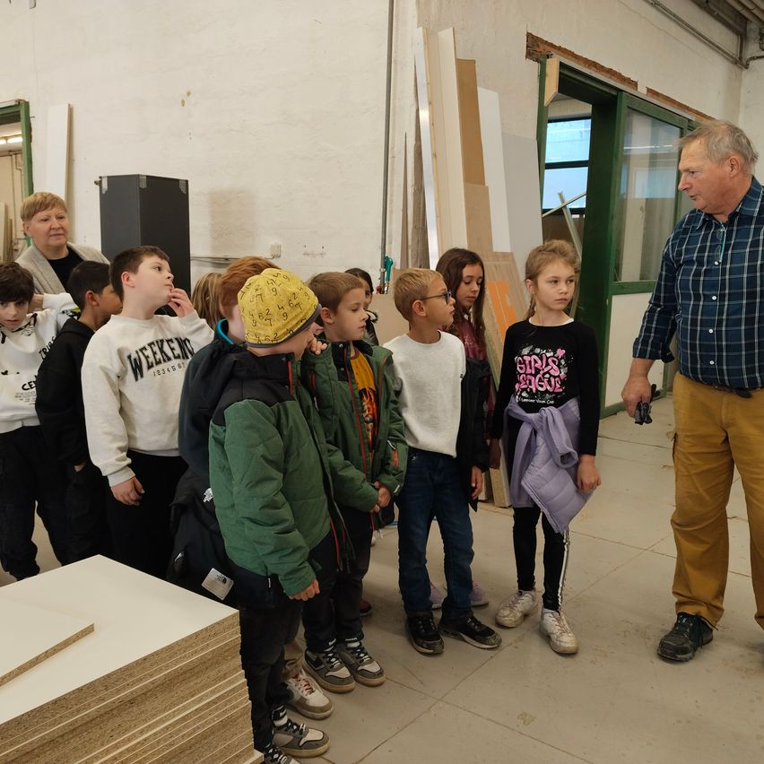 A group of children is standing in a room, looking at wooden boards. An adult man is explaining something to them.