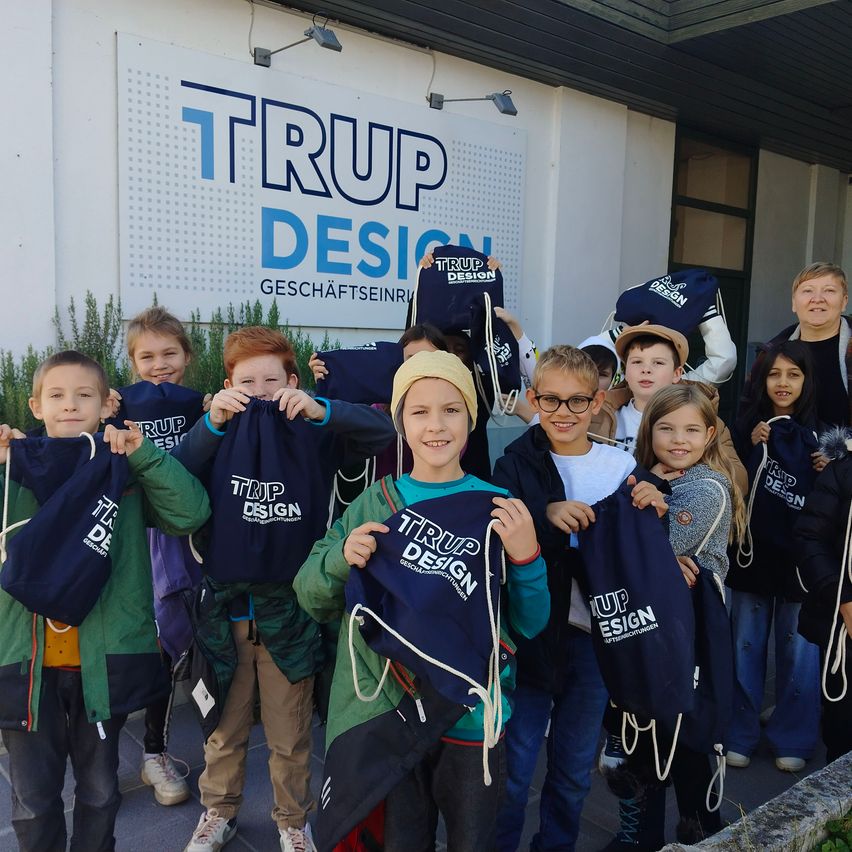 A group of children and adults stand outside in front of a building with a TRUP DESIGN sign. They are holding up blue bags with the TRUP DESIGN logo.