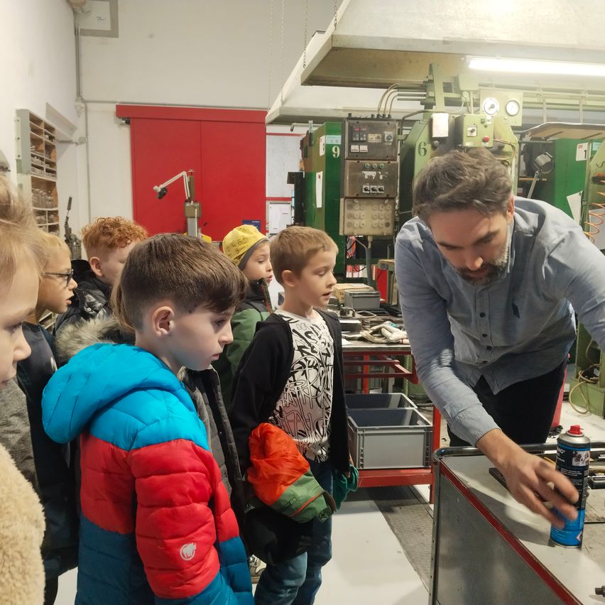 Ein Mann in einer Werkstatt erklärt etwas einer Gruppe von Kindern, die aufmerksam zuhören. Die Werkstatt ist mit Maschinen und Werkzeugen gefüllt.