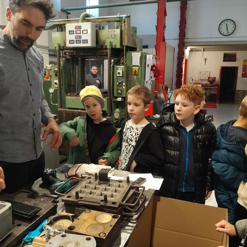 Ein Mann zeigt einer Gruppe von Kindern, wie man eine Maschine in einer Werkstatt benutzt. Die Kinder tragen Jacken und schauen sich die Maschine an.