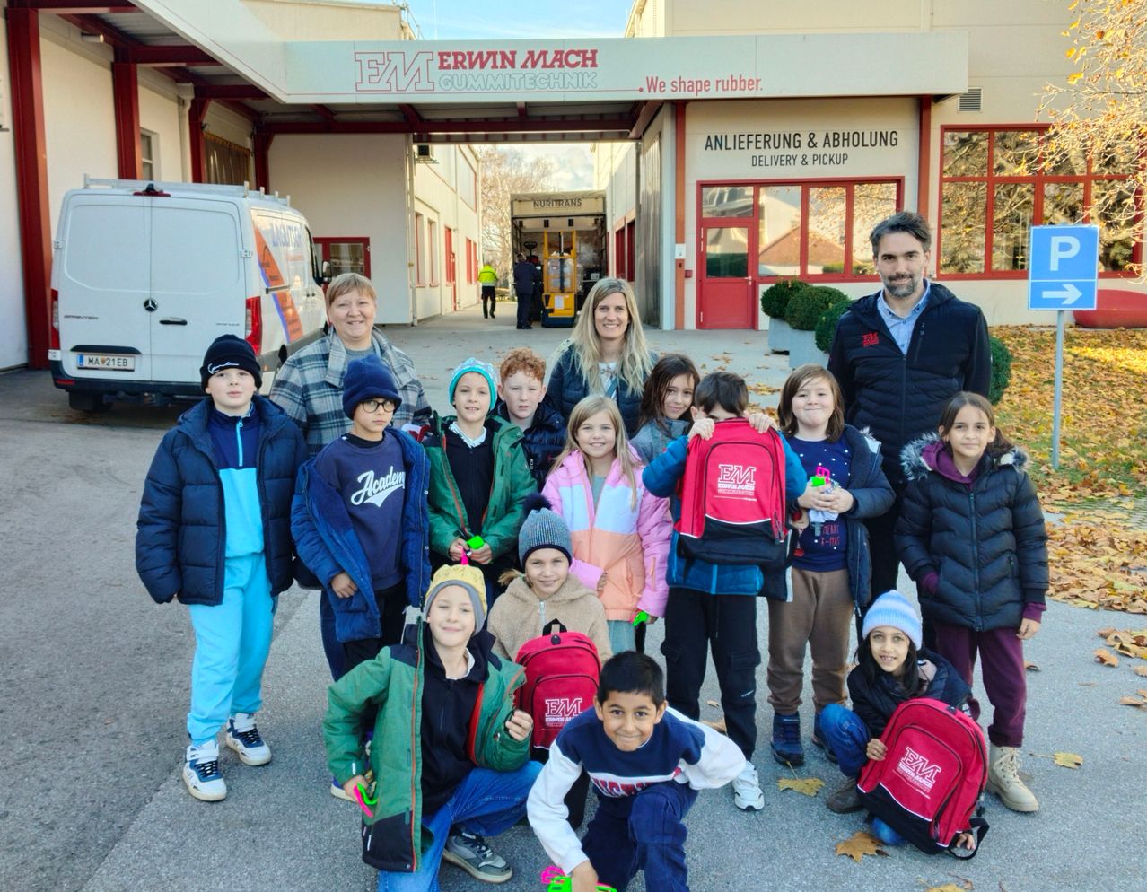 Eine Gruppe von Kindern mit Rucksäcken und zwei Erwachsenen posiert für ein Foto vor einem Gebäude mit dem Schild 'Erwin Mach Gummitechnik.' Ein weißer Van ist in der Nähe geparkt.