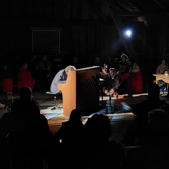 Ein Pianist spielt auf der Bühne in einem schwach beleuchteten Raum vor einem kleinen Publikum. Eine Lampe beleuchtet den Pianisten und eine Blumenarrangement in der Nähe.